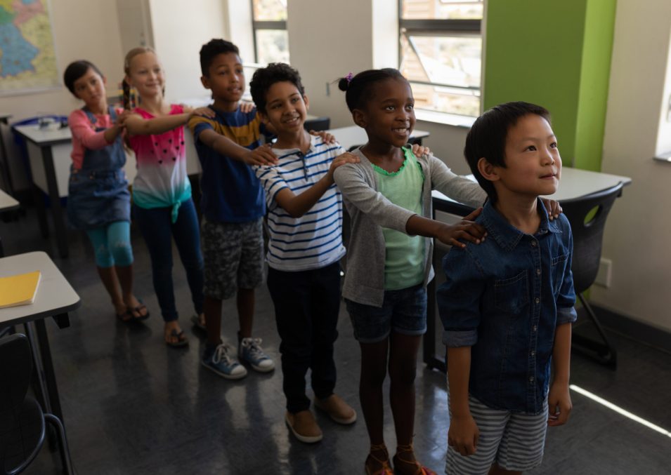 Side view of school kids standing in row with their hands on shoulder in classroom of elementary school